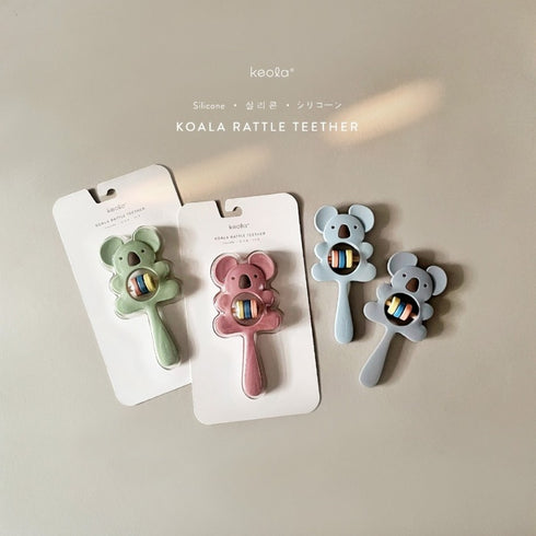 A set of three Koala Rattle Teethers in different colours packaged on white cardboard backing. Each teether features a bear-like design with a primary colour and a smaller secondary colour, and they all have a round rattle at the base.